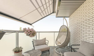 Cozy balcony seating area with a hanging wicker chair, beige cushions, a round table, and potted plants under a sunshade sunlit railing.
