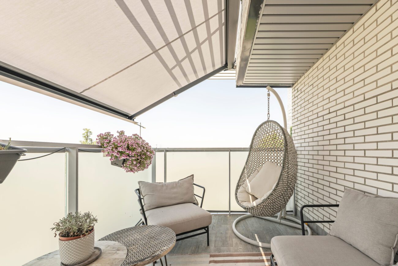 Cozy balcony seating area with a hanging wicker chair, beige cushions, a round table, and potted plants under a sunshade sunlit railing.