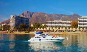 White luxury motor yacht on calm blue water near a sandy beach, with palm-lined promenade and multi-story hotels against rugged mountains behind them.
