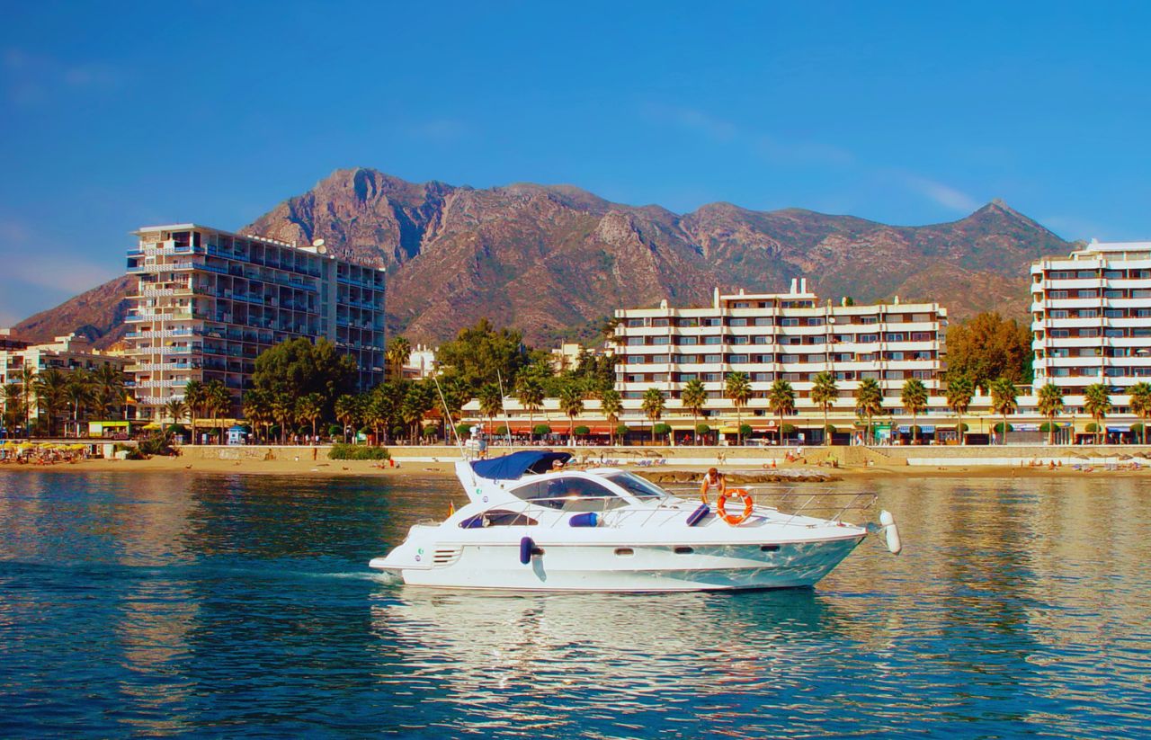 White luxury motor yacht on calm blue water near a sandy beach, with palm-lined promenade and multi-story hotels against rugged mountains behind them.