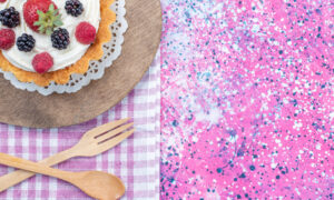 A berry-topped cake on a wooden board with a doily, beside a pink splatter-painted background and a purple checkered napkin with wooden cutlery nearby.