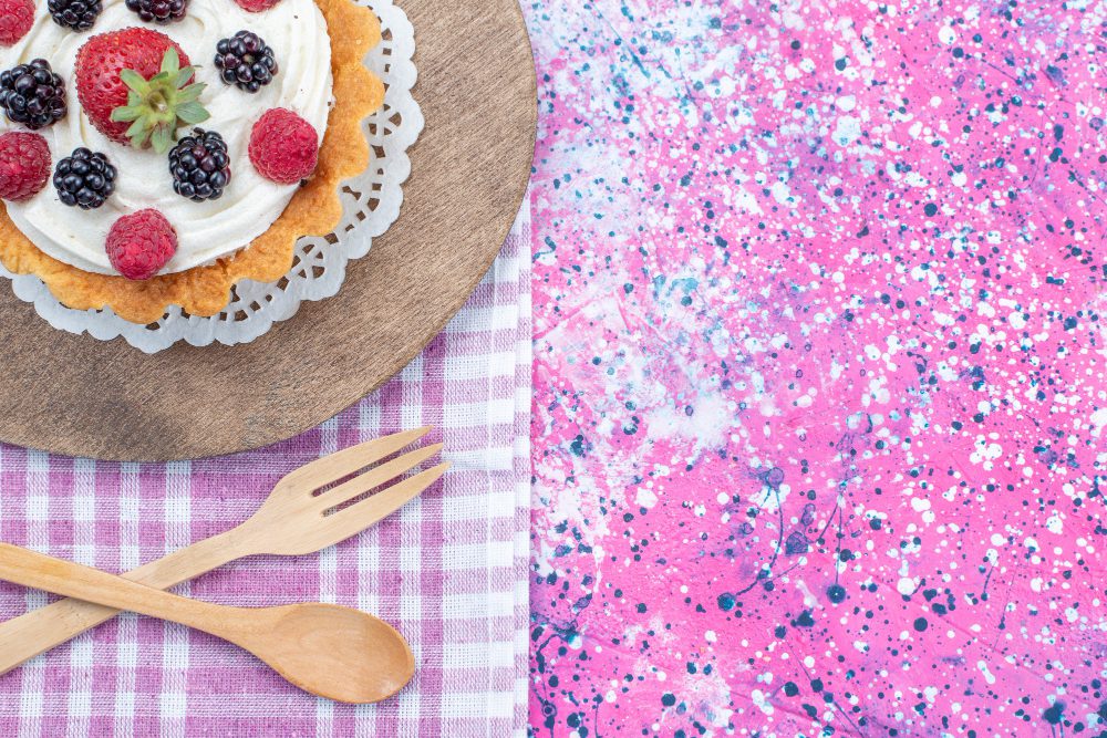 A berry-topped cake on a wooden board with a doily, beside a pink splatter-painted background and a purple checkered napkin with wooden cutlery nearby.