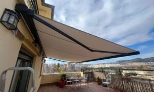 Terrace with a beige retractable awning extended, metal table and chairs, potted plants, overlooking a city and hills in the distance.