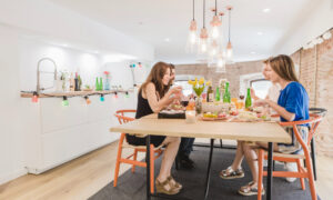 Group of five friends sharing a meal around a wooden table in a bright kitchen with bottles and string lights in the background.