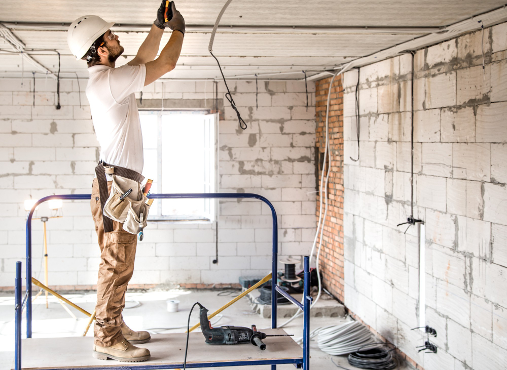 Construction worker wearing a hard hat on a scaffold, installing wiring to the ceiling in a renovation site.