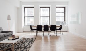 Bright minimalist living and dining space with a long wooden table and eight black chairs by three large windows; gray sofa, patterned rug, and floor lamp on light wood floors.