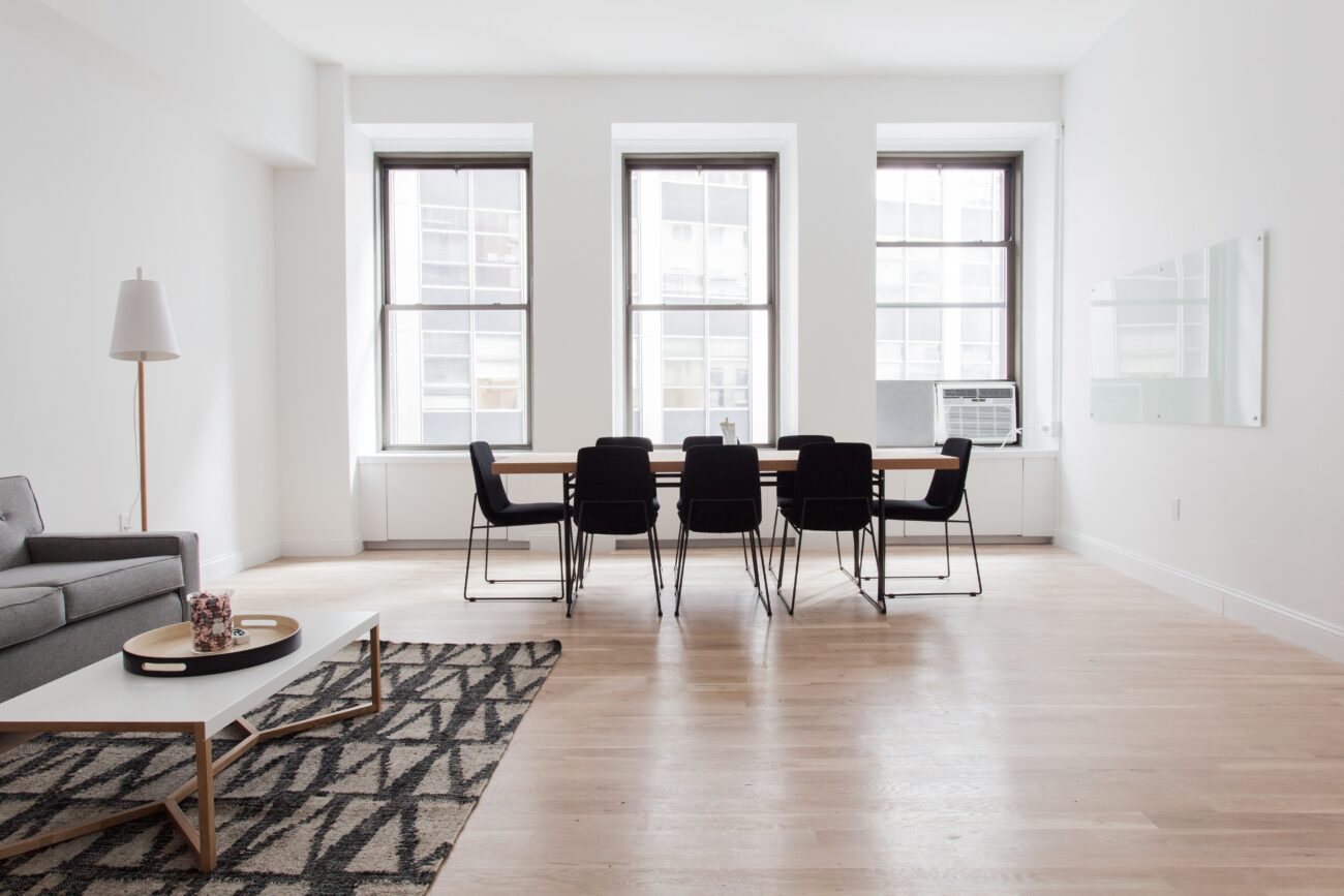 Bright minimalist living and dining space with a long wooden table and eight black chairs by three large windows; gray sofa, patterned rug, and floor lamp on light wood floors.
