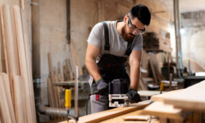 Carpenter in safety goggles using a router on a wooden plank in a workshop