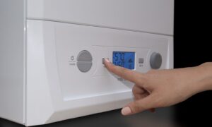 Person taps a button on a white boiler thermostat panel with a blue digital display showing settings and temperature.