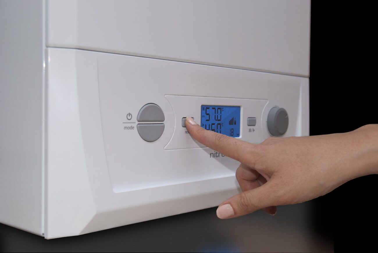 Person taps a button on a white boiler thermostat panel with a blue digital display showing settings and temperature.