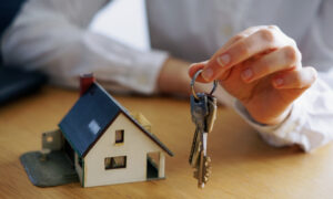 Hand holding a keyring with multiple house keys above a small model house on a wooden table.