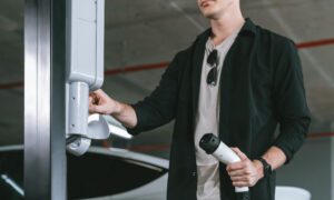 Man wearing a black shirt and white tee holds an EV charging plug near an electric car at a parking garage, about to connect it.