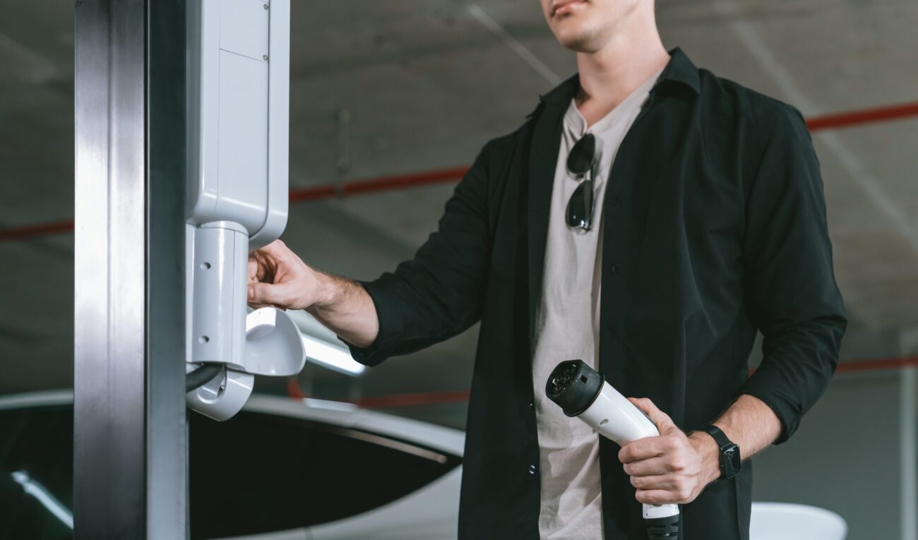 Man wearing a black shirt and white tee holds an EV charging plug near an electric car at a parking garage, about to connect it.