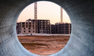 Construction site viewed through a circular concrete pipe opening; unfinished multi-story buildings and cranes in warm sunset light, bare dirt foreground