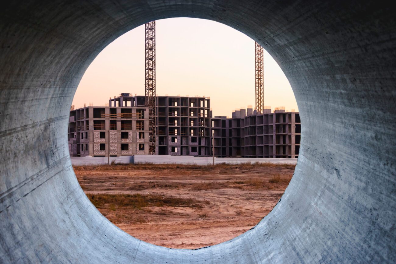 Construction site viewed through a circular concrete pipe opening; unfinished multi-story buildings and cranes in warm sunset light, bare dirt foreground