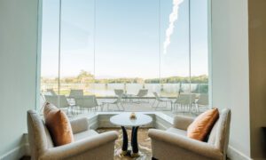 Two beige armchairs with orange pillows sit around a round white table by a floor-to-ceiling glass wall, overlooking an outdoor patio and water beyond.