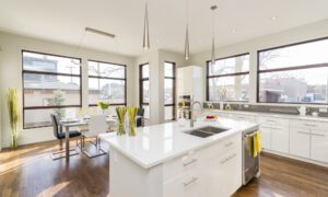 Bright, modern kitchen with white cabinets and an island, stainless sink, and a dining area by large windows to the left and back.