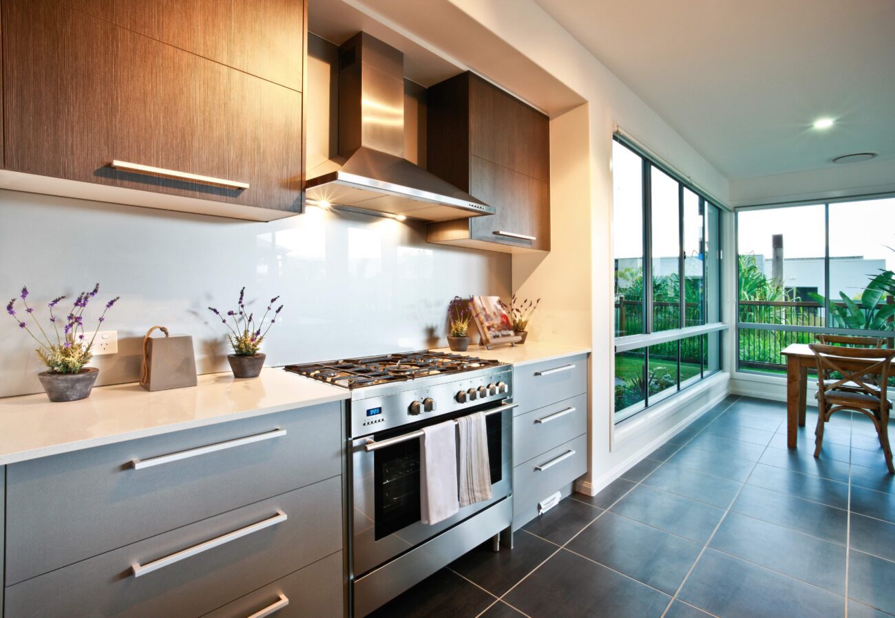 Modern kitchen with stainless steel range, hood, and gray drawers, lit by under-cabinet lights and large windows nearby.
