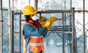 Construction worker in a yellow hard hat and orange high-visibility vest tightens scaffolding on a construction site, wearing gloves and a respirator mask.
