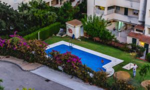 Rectangular blue swimming pool in a courtyard surrounded by flowering bushes and multi-story white apartment buildings.